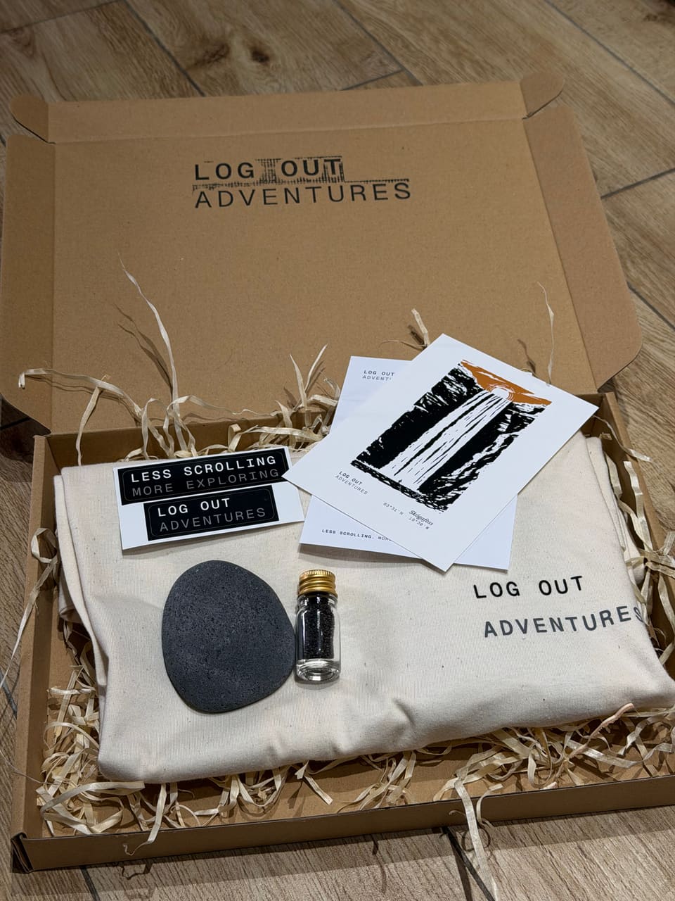 Pudełko LOG OUT Adventures z koszulką z logo, pocztówkami, naklejkami, kamieniem lawowym i fiolką czarnego piasku z Islandii